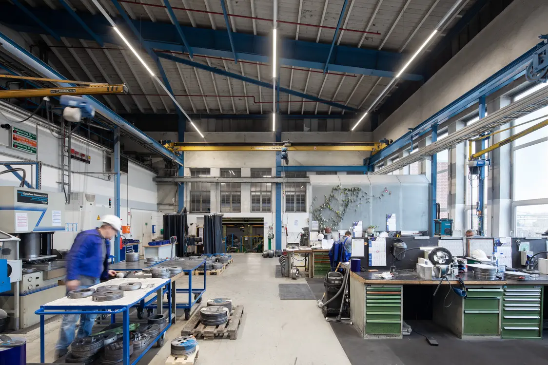 Eine helle Industriehalle mit hohen Decken, Werkbänken und Maschinen, an denen mehrere Arbeiter beschäftigt sind. Im Vordergrund arbeitet ein Mann mit Schutzhelm an einem Tisch, während im Hintergrund weitere Arbeitsplätze und Geräte zu sehen sind.