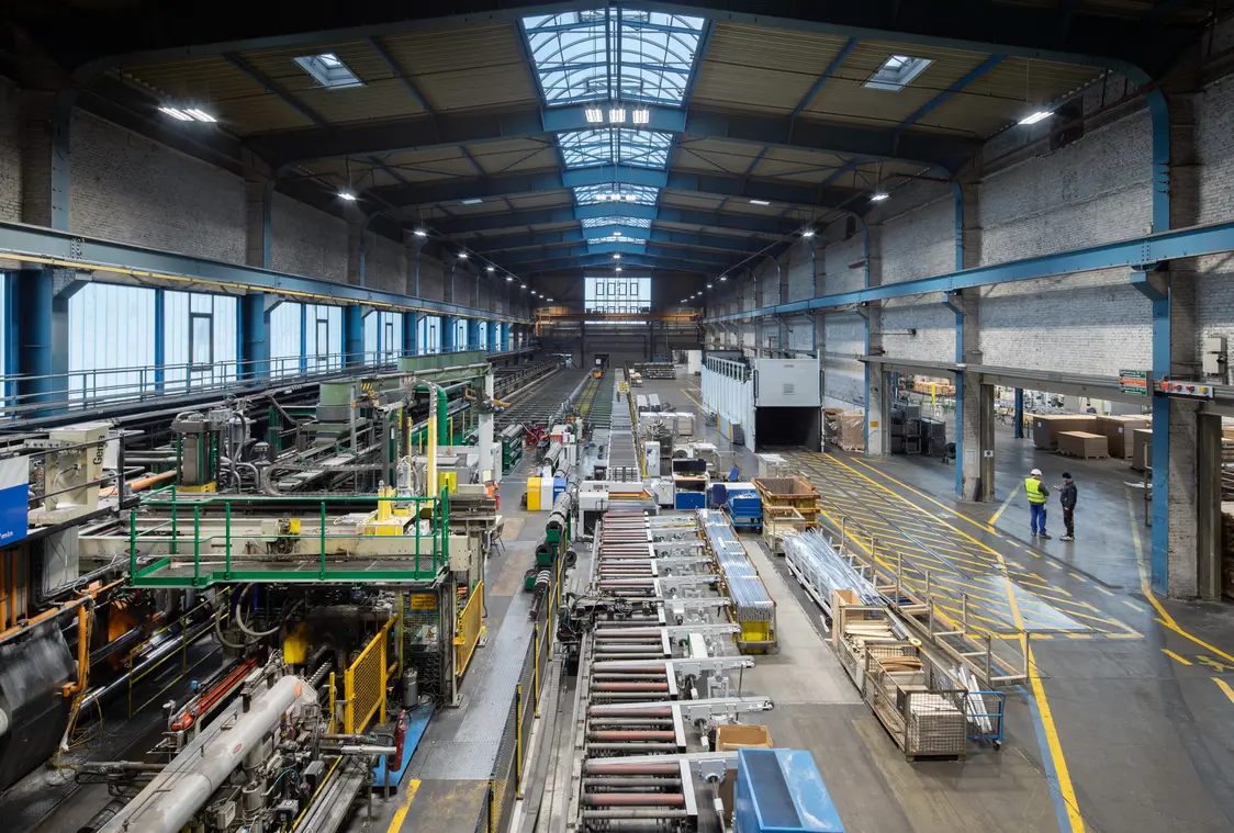 Eine große, helle Industriehalle mit Maschinen, Förderbändern und Arbeitsbereichen. Im Hintergrund laufen mehrere Personen durch die Halle, während im Vordergrund Produktionsanlagen zu sehen sind.