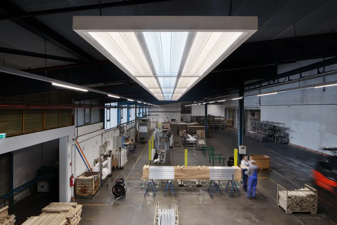 Eine große, helle Industriehalle mit mehreren Werkbänken und Maschinen, in der ein Arbeiter an einem Tisch arbeitet. Im Vordergrund sind Metallprofile und Arbeitsmaterialien zu sehen.