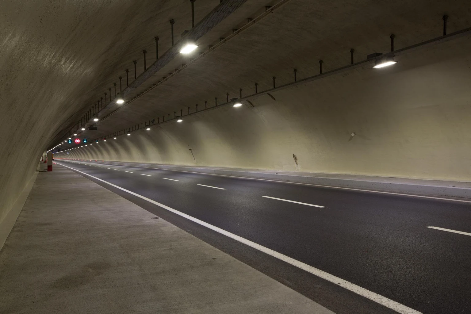 Un tunnel routier spacieux, aux parois en béton lisse et à la chaussée proprement asphaltée. Le tunnel est éclairé par des luminaires montés au plafond, régulièrement espacés, et les marquages au sol assurent une parfaite lisibilité de la voie pour les automobilistes.