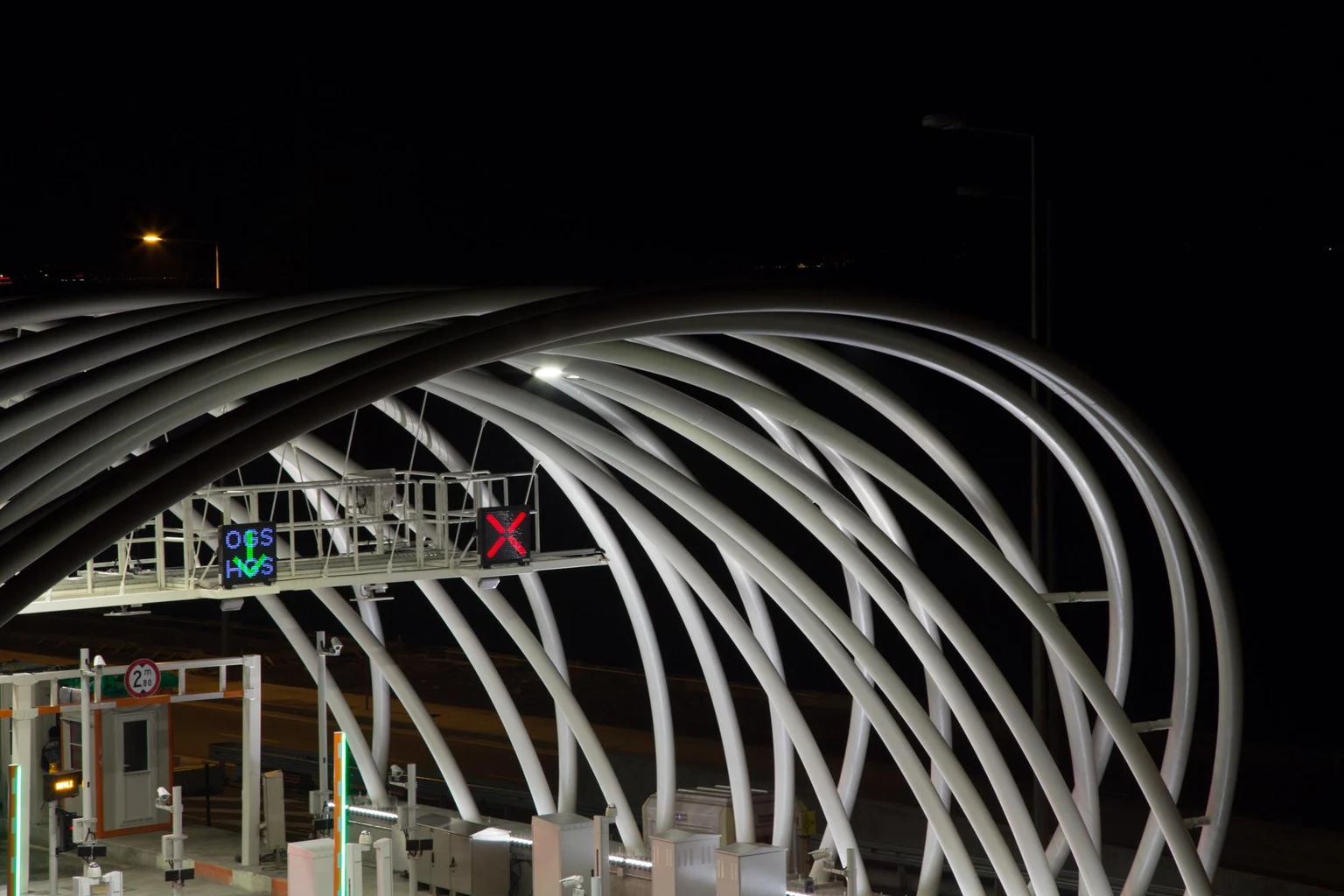 Un poste de péage moderne éclairé de nuit, avec des structures métalliques arquées et un éclairage LED intense. La station est éclairée par des lumières puissantes, et les voies de circulation sont équipées de panneaux électroniques, incluant des flèches vertes et des croix rouges pour réguler le flux de véhicules. Le style architectural épuré apporte une touche contemporaine à cette infrastructure fonctionnelle.
