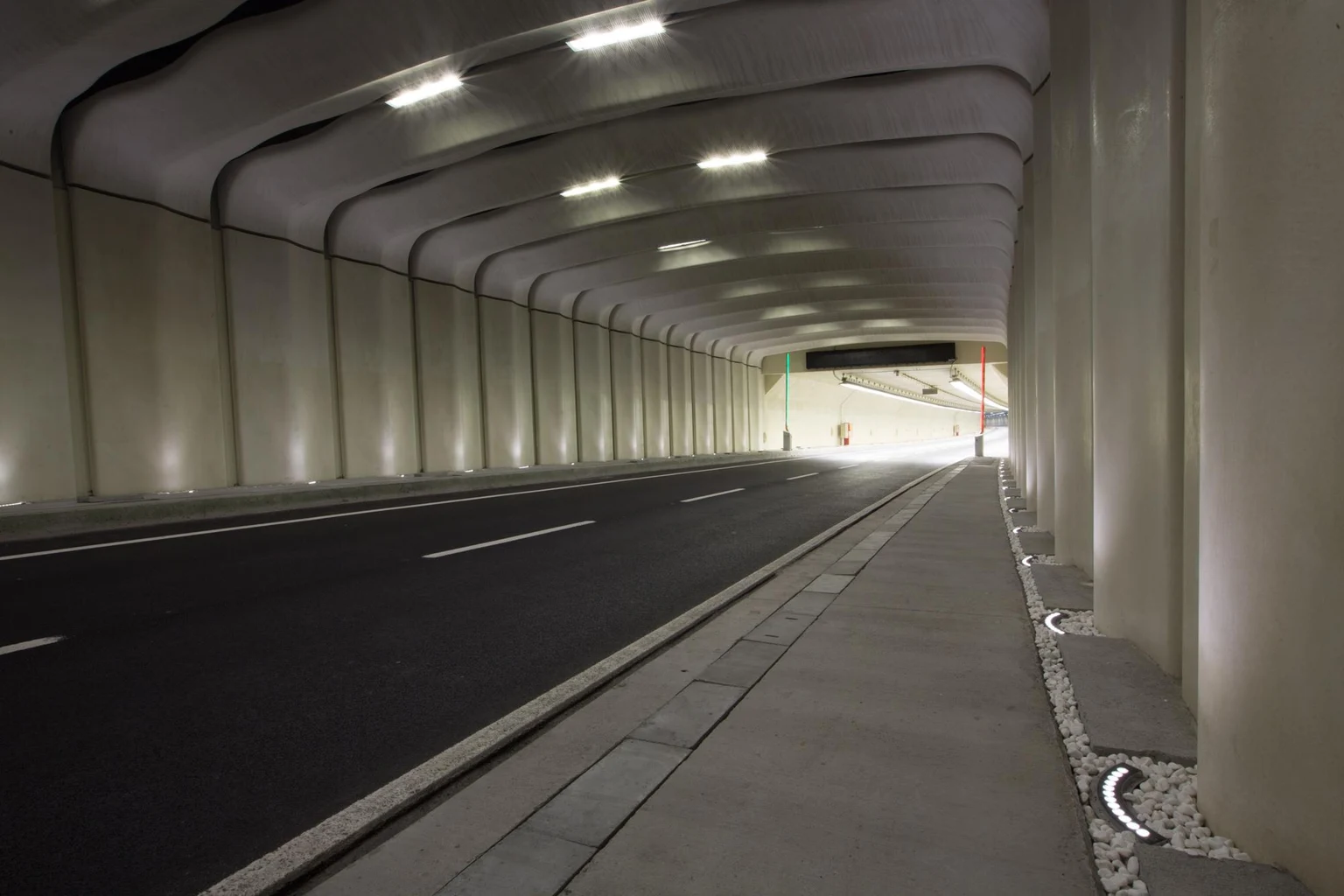  Un tunnel routier bien éclairé aux parois incurvées, avec une sortie visible au loin. L’espace est équipé de luminaires LED montés au plafond, de plots réfléchissants sur la chaussée et d’un passage piéton latéral pour plus de sécurité.