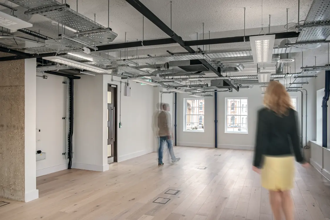 Modern office space with large windows and ample daylight, open ceiling featuring technical installations and linear ceiling luminaires as a lighting solution.