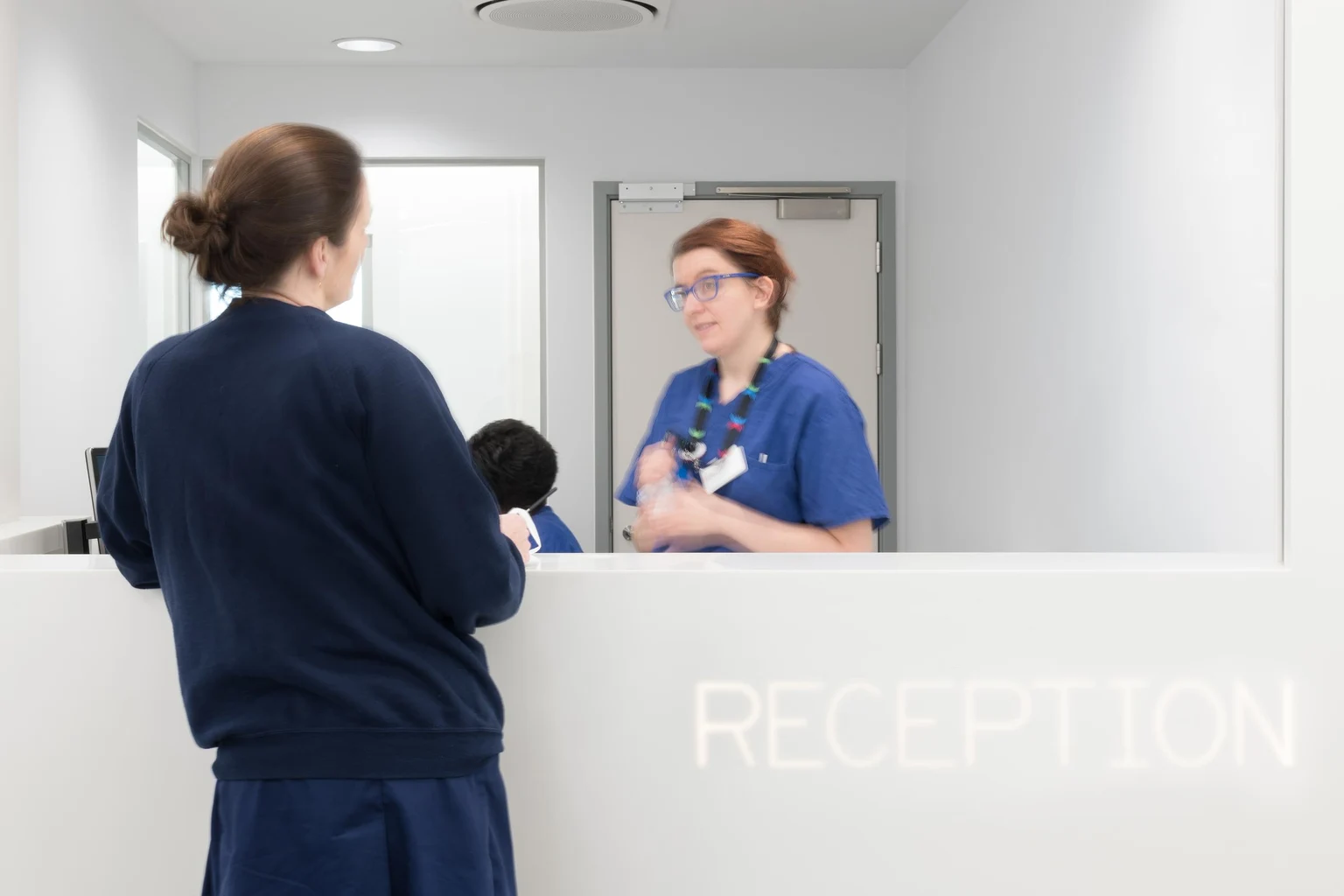 Een vrouw in een blauw uniform staat aan een witte balie te praten met een andere vrouw in blauwe operatiekleding. Een led-downlight op de achtergrond verlicht de receptie.