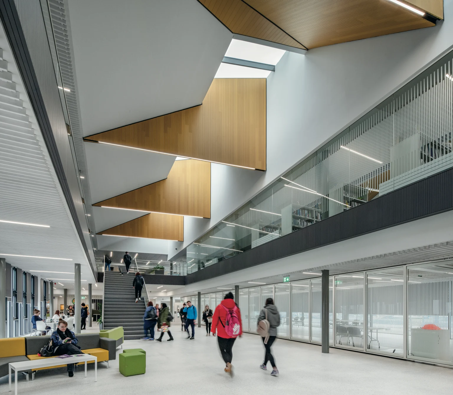Forth Valley College, Falkirk - People walk through a modern,  bright and welcoming reception area, with a high ceiling featuring recessed seamlessly integrated lighting, cushioned seating to the left hand-side and a staircase in the background