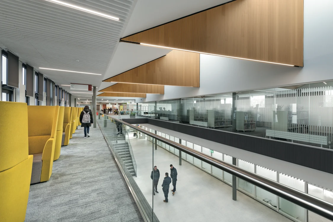 Forth valley College, Falkirk - a person walks along a balcony lined with yellow, cushioned seating, a staircase leading down in the background, where 3 people stand on the ground floor