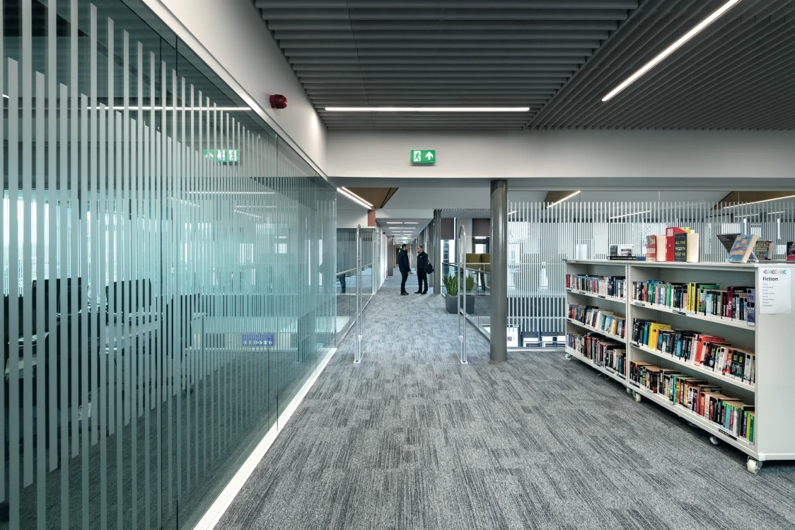 Forth Valley College, Falkirk - A corridor with a frosted glass partition running along side it, featuring shelves filled with books to the right and 2 people in the background