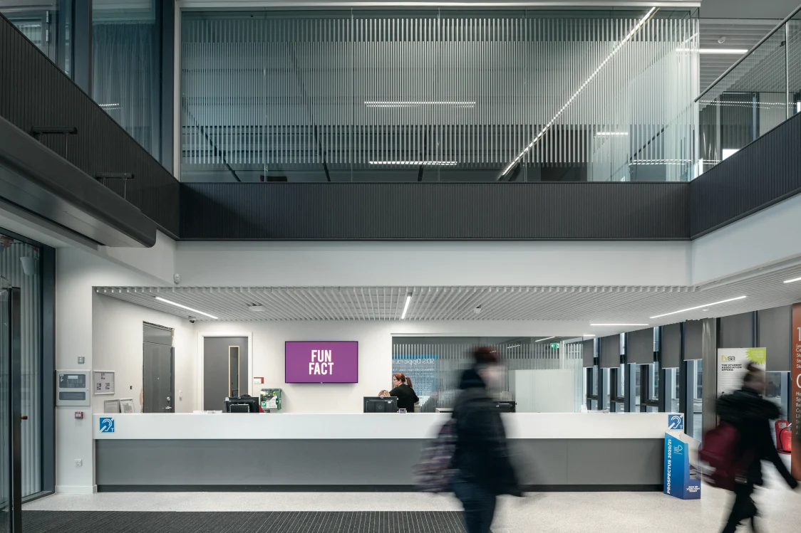 Forth Valley College, Falkirk - A modern reception desk with a screen mounted on the wall and 2 computers. A lady stands behind the desk, whilst 2 people walk past