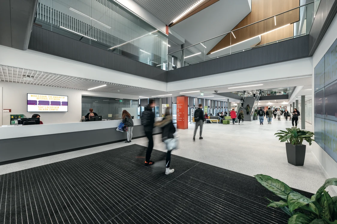 Forth Valley College, Falkirk - People walk through a modern,  bright and welcoming reception area, with a high ceiling and recessed seamlessly integrated lighting and a staircase and seating in the background. 1 lady stands at the desk whist 1 person stands and 1 sits behind the desk