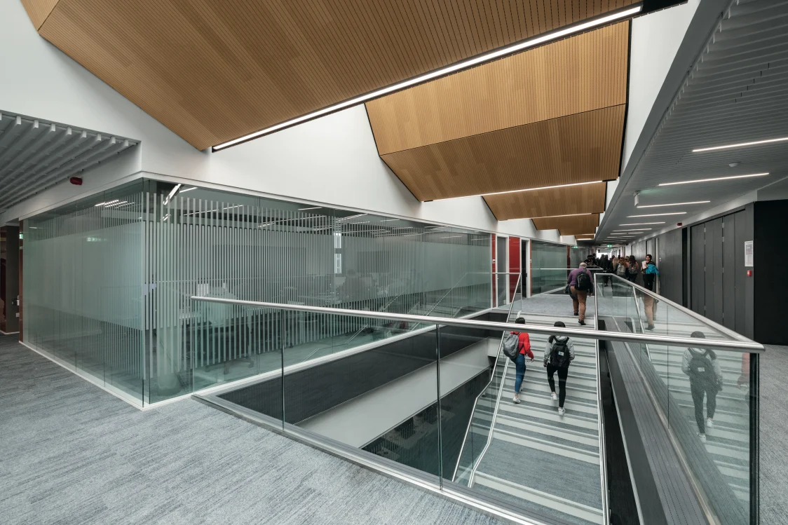 Forth valley College, Falkirk - 2 people walk down a modern staircase located next to a glass frosted partition with a balcony in the foreground