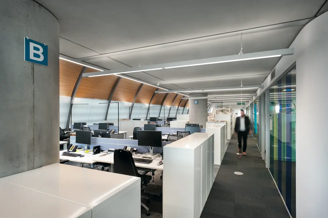 Längliche Deckenleuchten sorgen für eine helle, gleichmäßige Ausleuchtung des Flurs und der Arbeitsplätze; Tageslicht fällt durch die Fensterfront ein.