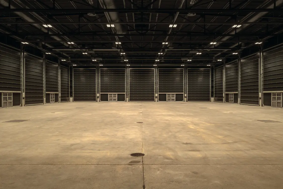 Große, leere Industriehalle mit Betonboden und hoher Decke, beleuchtet durch zahlreiche Deckenleuchten. Die Wände sind dunkel und es sind keine Einbauten oder Maschinen zu sehen.