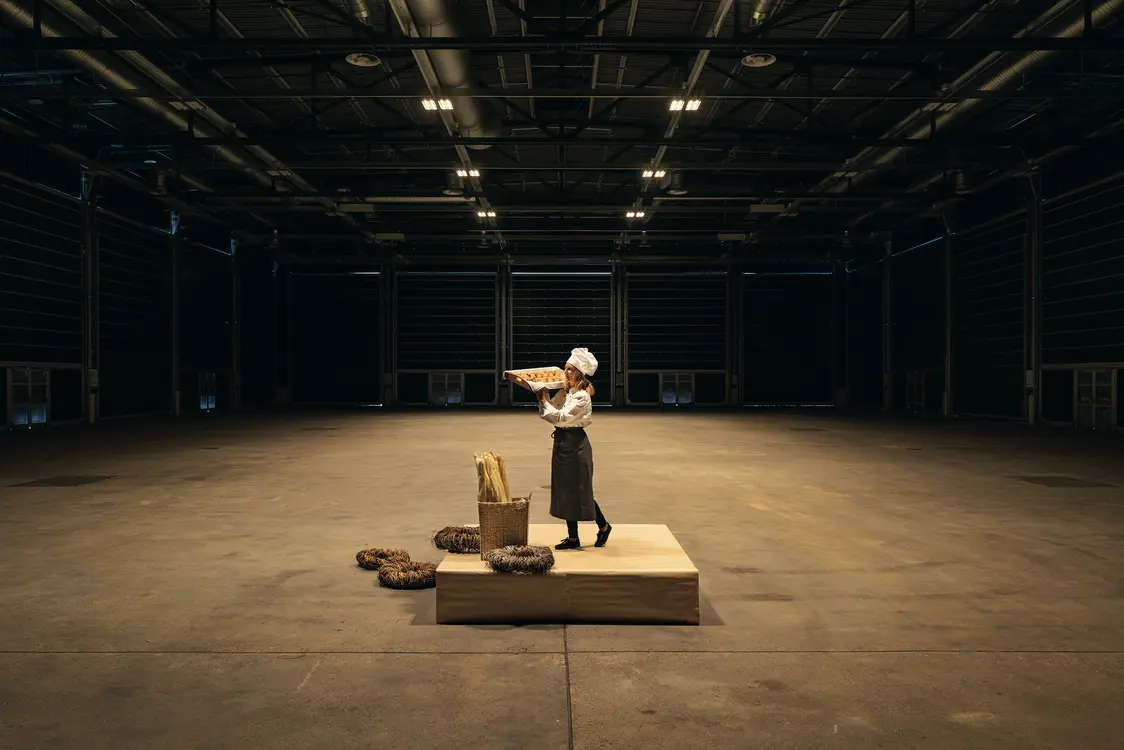 Eine Person in Bäckeruniform steht auf einem Podest in einer großen, leeren Industriehalle und hält ein Brot in den Händen. Um sie herum liegen weitere Brote und eine Holzkiste auf dem Boden.