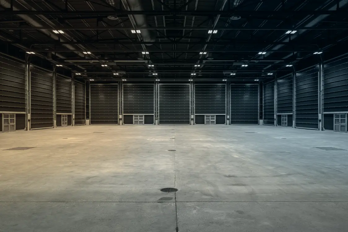 Große, leere Industriehalle mit Betonboden und hoher Decke, beleuchtet durch zahlreiche Deckenleuchten. Die Wände sind dunkel und es sind keine Maschinen oder Einbauten vorhanden.