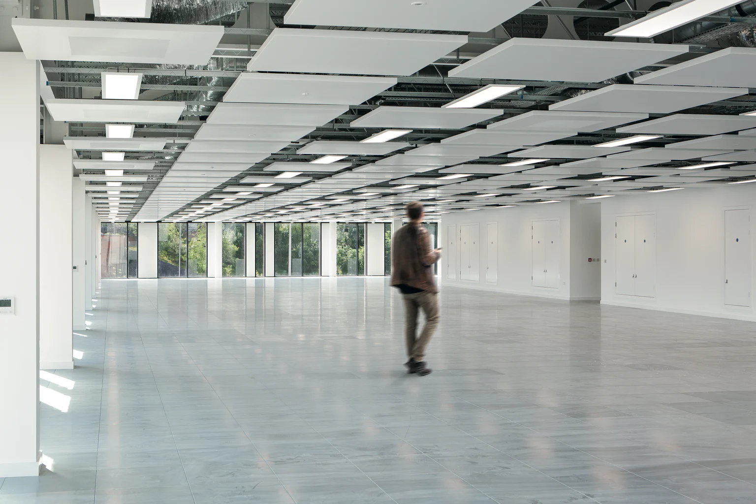 Eine verschwommene Gestalt bewegt sich durch eine geräumige, helle und minimalistische Büroumgebung mit weißen Wänden und großen Fenstern auf einer Seite.