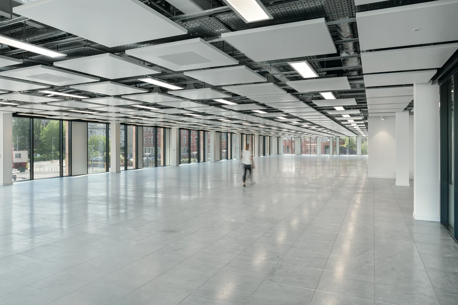 Eine Person geht durch einen weitläufigen, offenen Bürobereich mit spiegelnden Böden, hohen Decken und großen Glaswänden, die den Blick nach draußen freigeben.