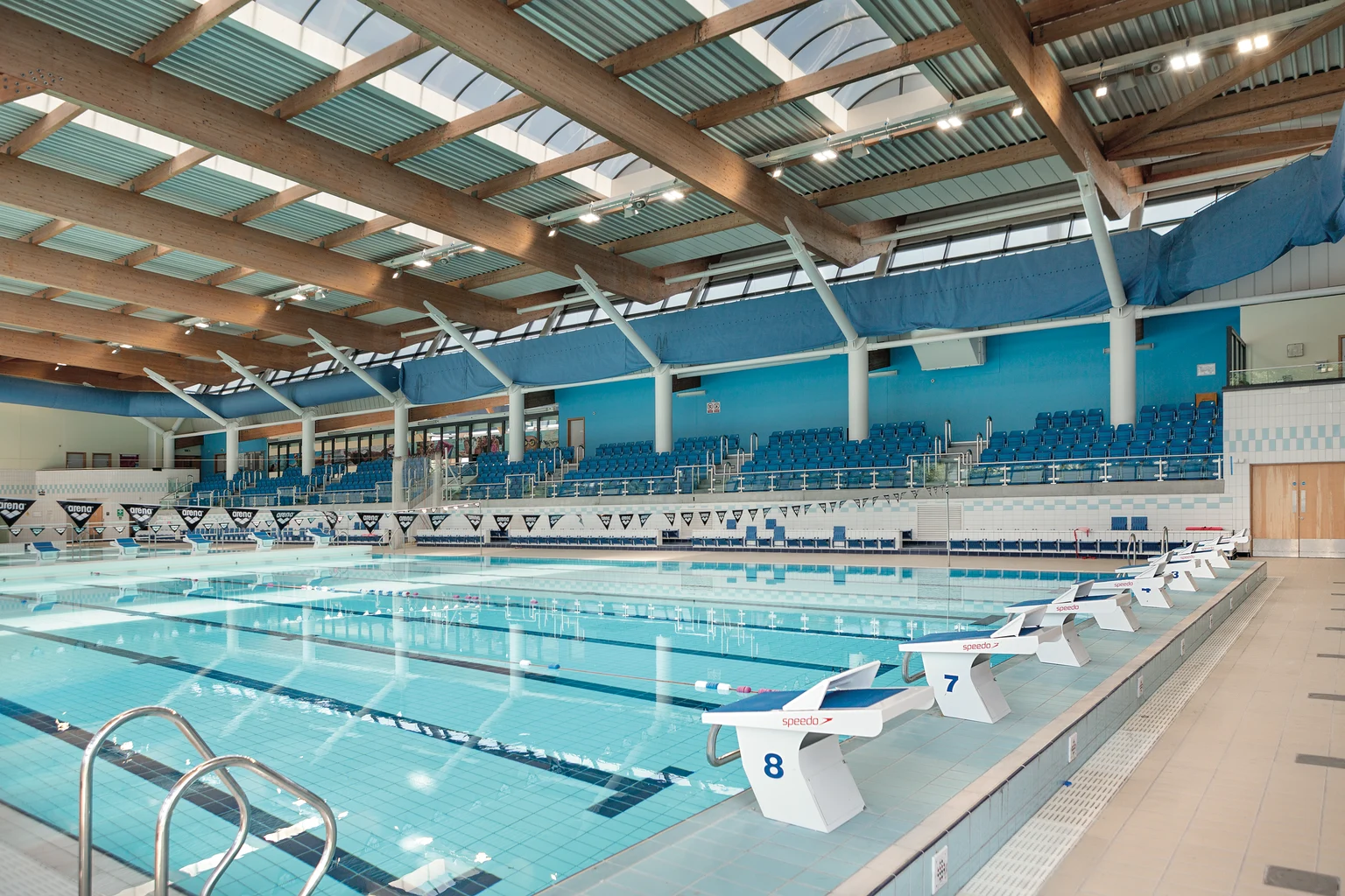 Het interieur van het overdekte zwembad van het Liverpool Aquatics Centre met meerdere banen met startblokken. Houten balken, blauwe zitgedeelten en grote vensters creëren een lichte en ruimtelijke sfeer.