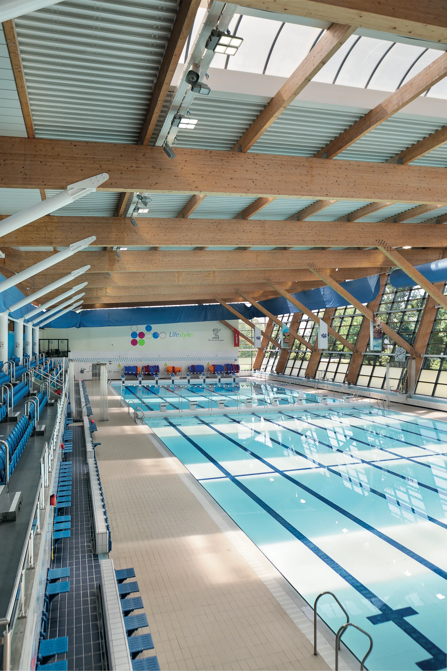 Het interieur van een modern binnenzwembad met meerdere banen, omgeven door blauwe zitgedeelten. Houten balken en grote vensters zorgen voor natuurlijk licht in de ruimte, waardoor er een lichte en open sfeer ontstaat. De kwaliteit van de verlichting verbetert de veiligheid, zichtbaarheid en algehele ervaring voor zwemmers en bezoekers.