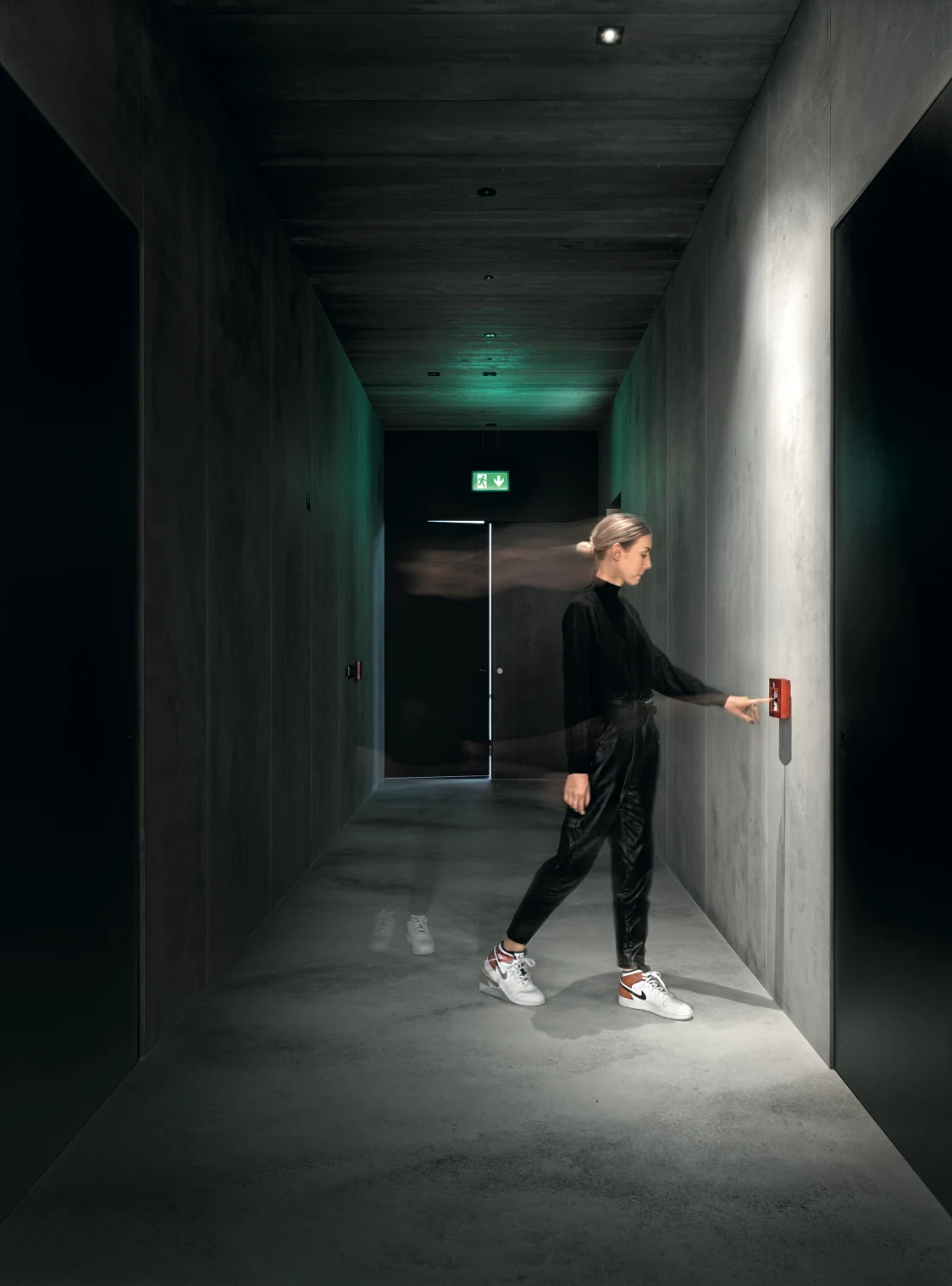 A modern concrete corridor with a well-lit emergency exit sign next to an escape door.