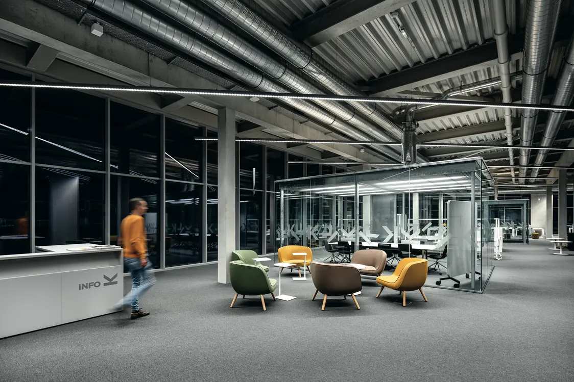An open office space featuring an industrial design, exposed pipes, and glass partitions. Linear LED luminaires ensure uniform illumination, while colorful seating furniture creates an inviting atmosphere.