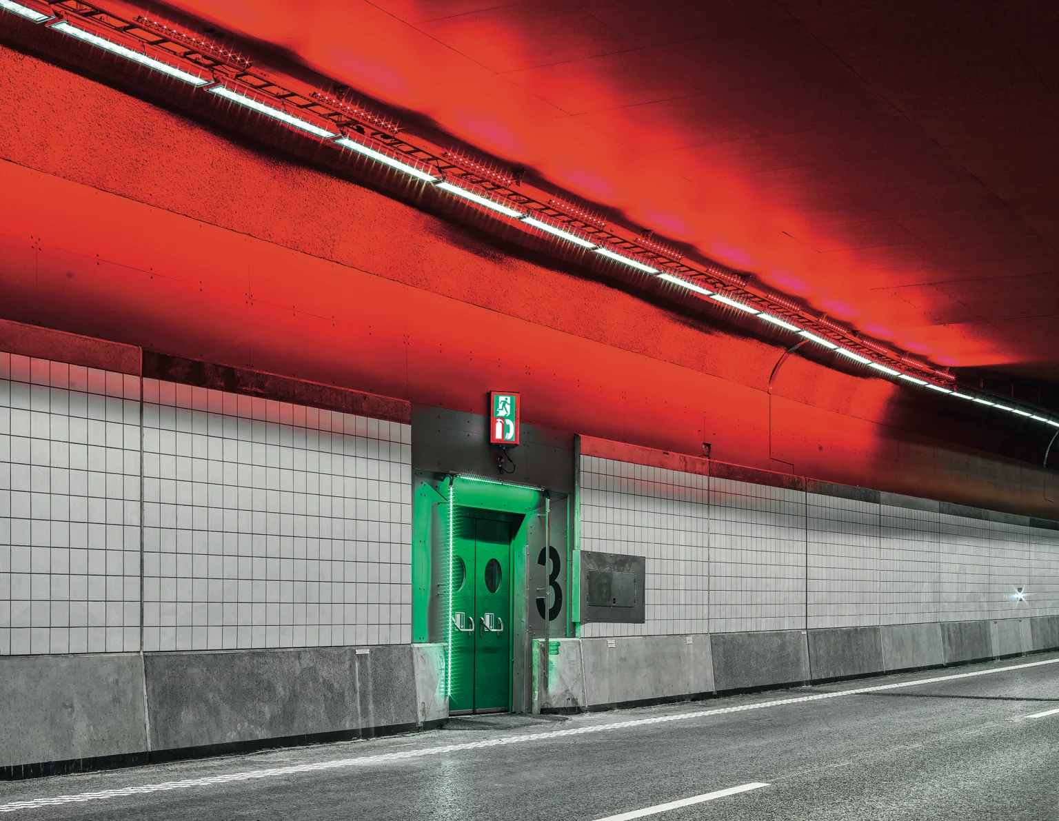 Een moderne verkeerstunnel met een opvallend rood plafond, verlicht door lineaire verlichting. In de betegelde muren zit een groene nooduitgangsdeur, die een fel contrast vormt met de neutrale tinten eromheen.