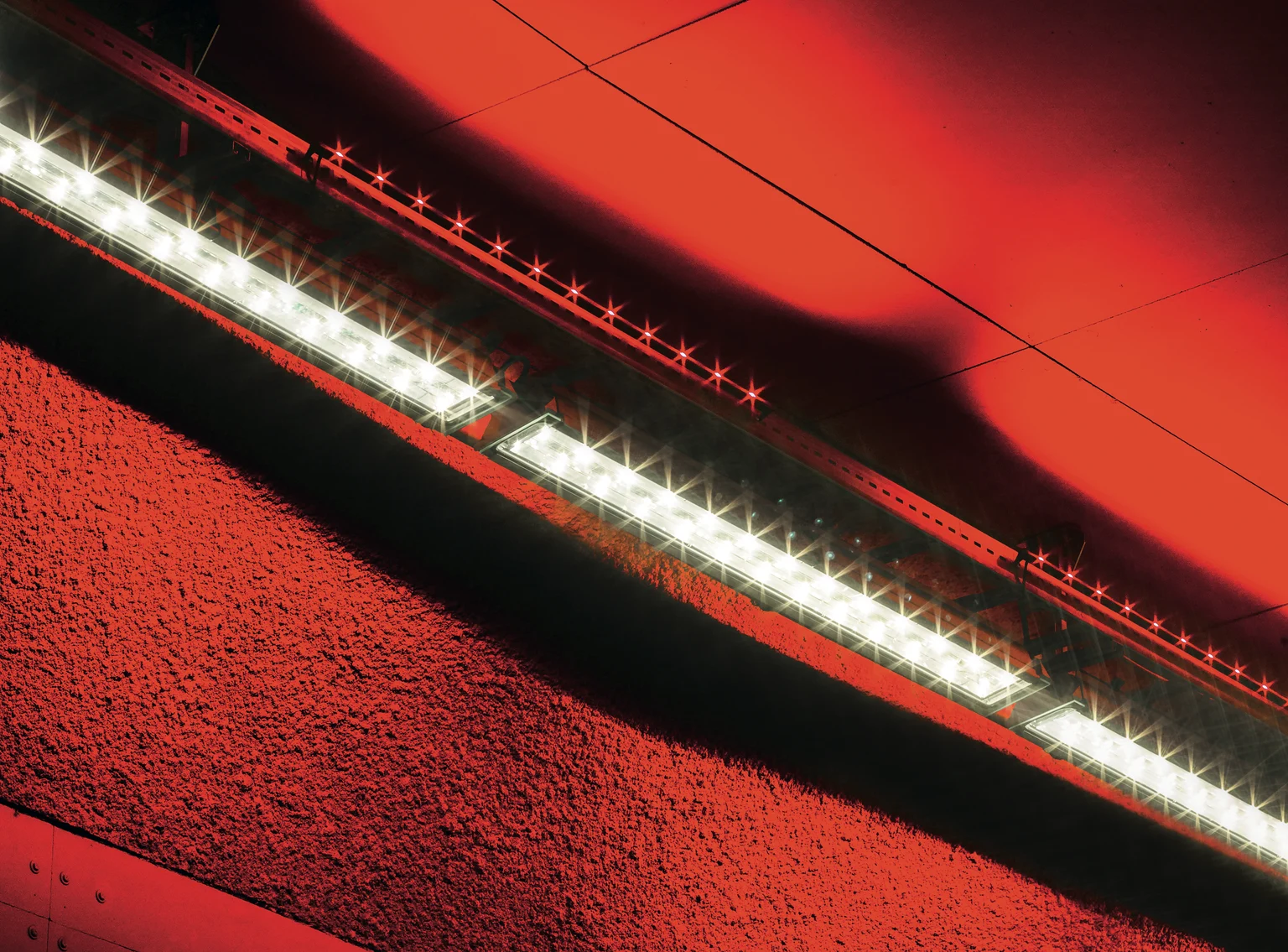 Een close-up van moderne, in een tunnel geïnstalleerde ledverlichting die helder wit licht uitstraalt. Het roodgeverfde plafond en de muur met structuur vormen een opvallend contrast met het strakke, lineaire ontwerp van de armaturen.