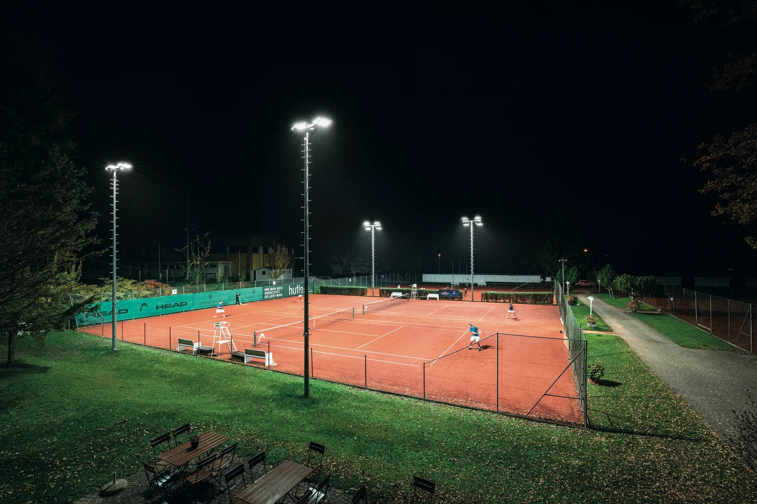 Un campo da tennis all'aperto ben illuminato di notte, circondato da recinzioni verdi e alberi. I giocatori sono visibili sulla superficie di terra rossa, illuminata da alti proiettori.