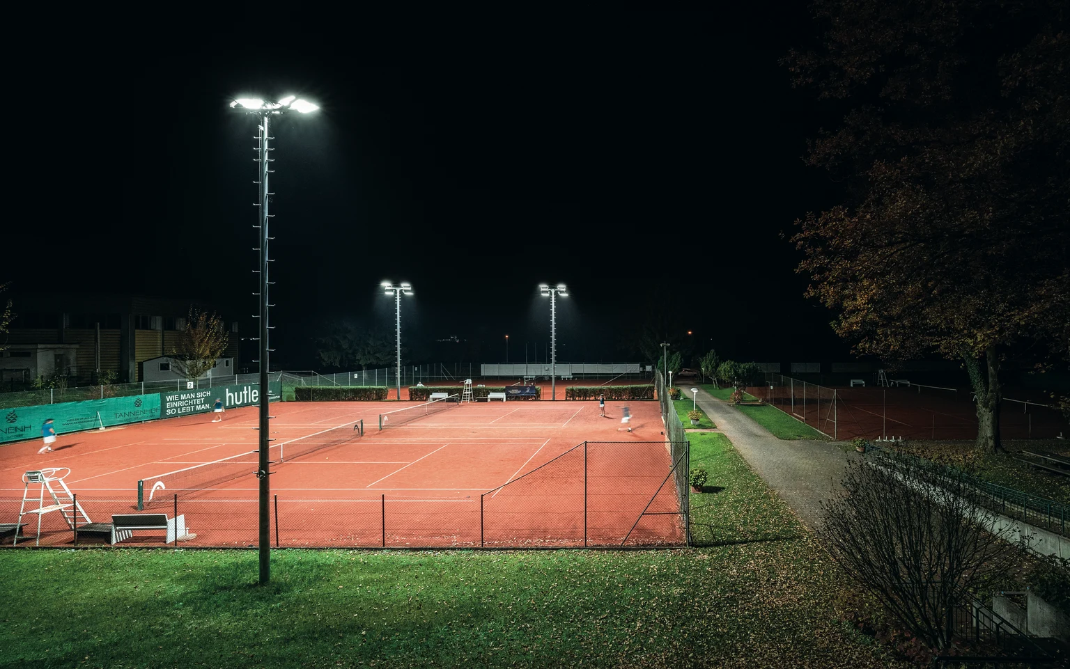 Un campo da tennis all'aperto ben illuminato di notte, circondato da recinzioni verdi e alberi. I giocatori sono visibili sulla superficie di terra rossa, illuminata da alti proiettori.