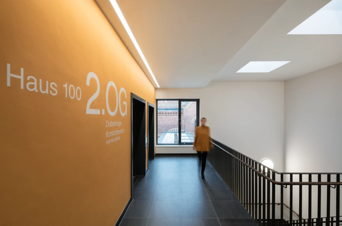 A bright corridor with an orange wall and linear LED lights on the ceiling. The lighting provides clear orientation and a welcoming atmosphere.