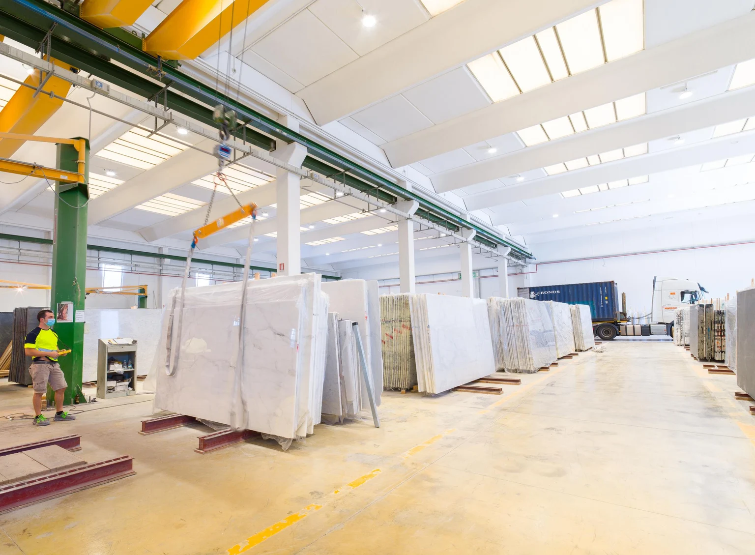 Un hall d’usine lumineux avec de grandes dalles de marbre soigneusement empilées sur des palettes. L’espace est bien éclairé par des puits de lumière et équipé de systèmes de grues pour la manutention des matériaux.