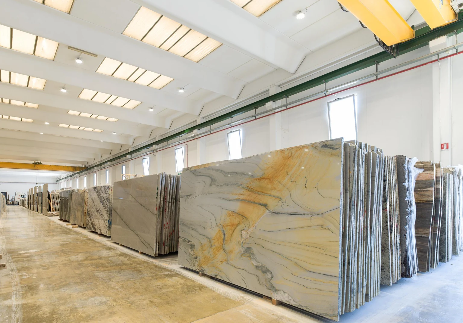 Un hall d’usine lumineux avec de grandes dalles de marbre soigneusement alignées en rangées. L’espace dispose de hauts plafonds, de puits de lumière et de systèmes de grues pour une manutention efficace des matériaux.