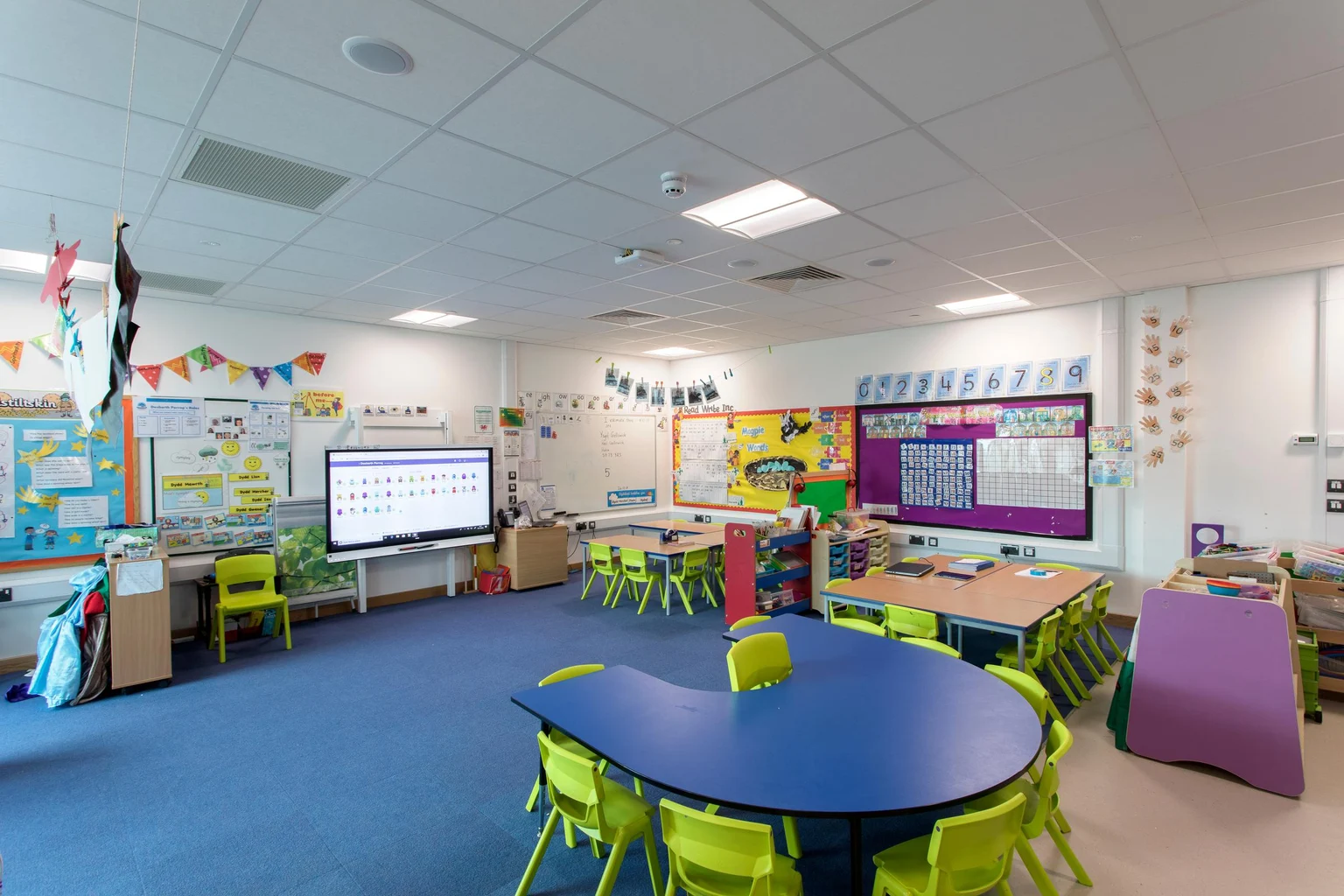 Een helder en kleurrijk klaslokaal met groene stoelen, een blauwe tafel en muren voorzien van educatief materiaal en kunstwerken van leerlingen. De ruimte bevat een interactief whiteboard en is goed verlicht met plafondverlichting.