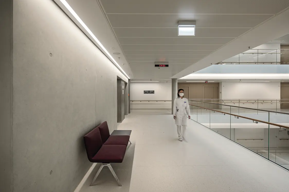 Ein heller Krankenhausflur mit minimalistischem Design. Die Decke ist mit integrierten LED-Leuchten ausgestattet, die für eine gleichmäßige Beleuchtung sorgen. Eine Person in weißer Kleidung geht entlang des Flurs, der durch Sitzgelegenheiten in Bordeauxrot und Glasgeländer ergänzt wird.