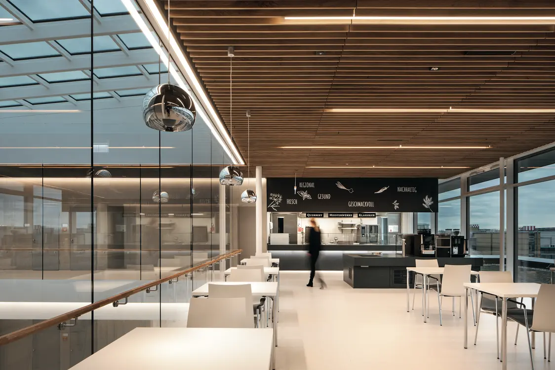 Ein moderner Gemeinschaftsbereich mit einer offenen Gestaltung und großen Glaswänden. Die Decke ist mit Holzlamellen und linearen LED-Leuchten ausgestattet, die für eine elegante und energieeffiziente Beleuchtung sorgen. Weiße Tische und Stühle schaffen eine klare und freundliche Atmosphäre, während im Hintergrund eine Person an der Kaffeestation sichtbar ist.