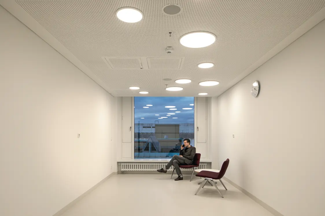 Ein heller, minimalistischer Raum mit weißen Wänden und Decken, ausgestattet mit runden LED-Einbauleuchten, die für eine gleichmäßige Beleuchtung sorgen. Im Hintergrund sitzt eine Person auf einem Stuhl vor einem großen Fenster, das Tageslicht hereinlässt und den Blick nach draußen freigibt.