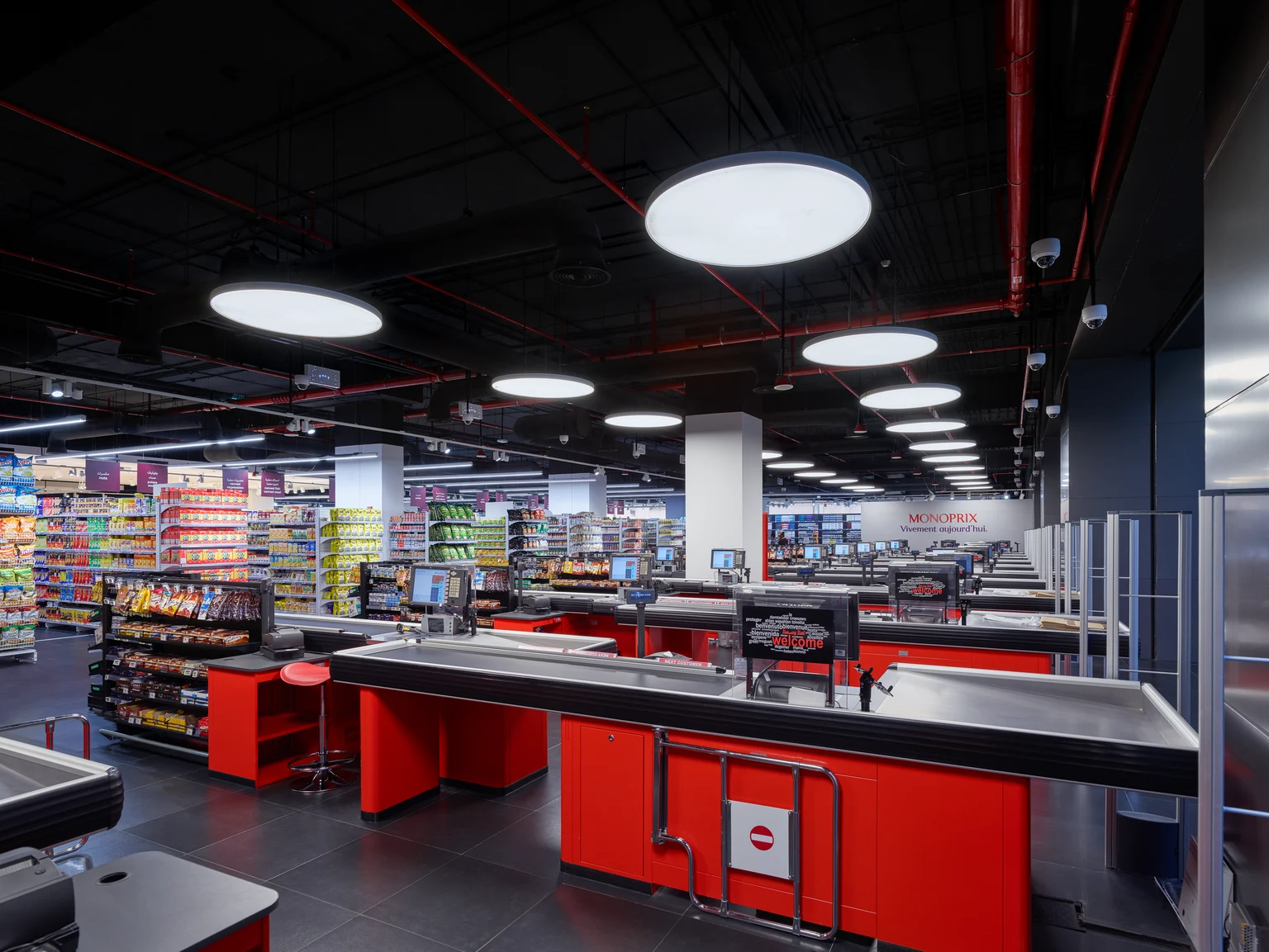 Une zone de passage en caisse spacieuse dans un supermarché avec des caisses rouges modernes, des stations en libre-service et des rayons remplis de produits en arrière-plan.