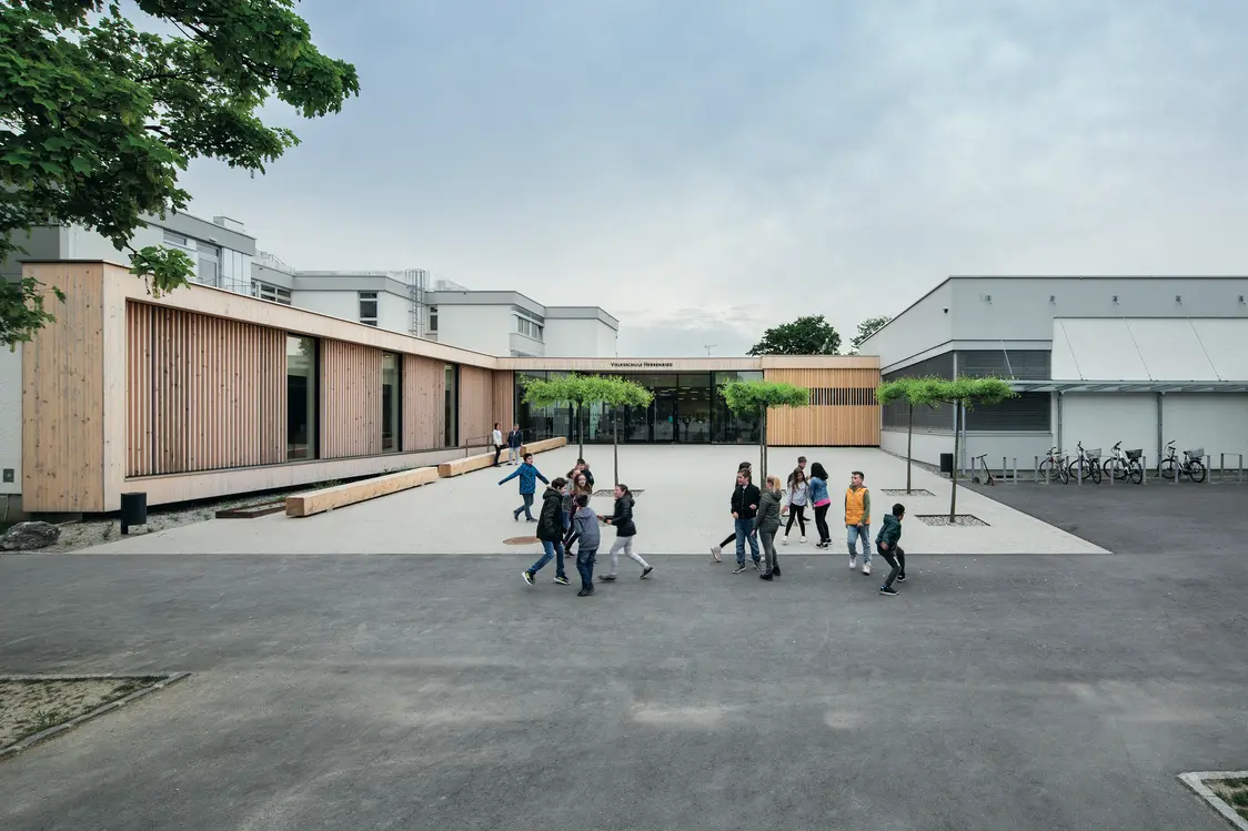 L'area esterna di una scuola con una moderna facciata in legno e un ampio parco giochi. I bambini giocano nel parco, mentre gli alberi e le panchine creano un ambiente piacevole.