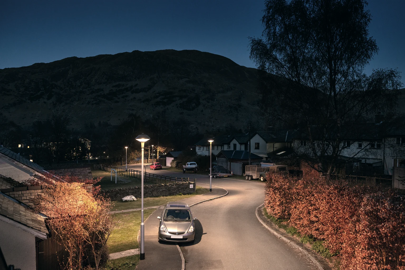 Een rustige straat in een woonwijk 's nachts met een enkele geparkeerde auto, verlicht door straatlantaarns. Huizen en bomen staan langs de weg, met donkere heuvels op de achtergrond.
