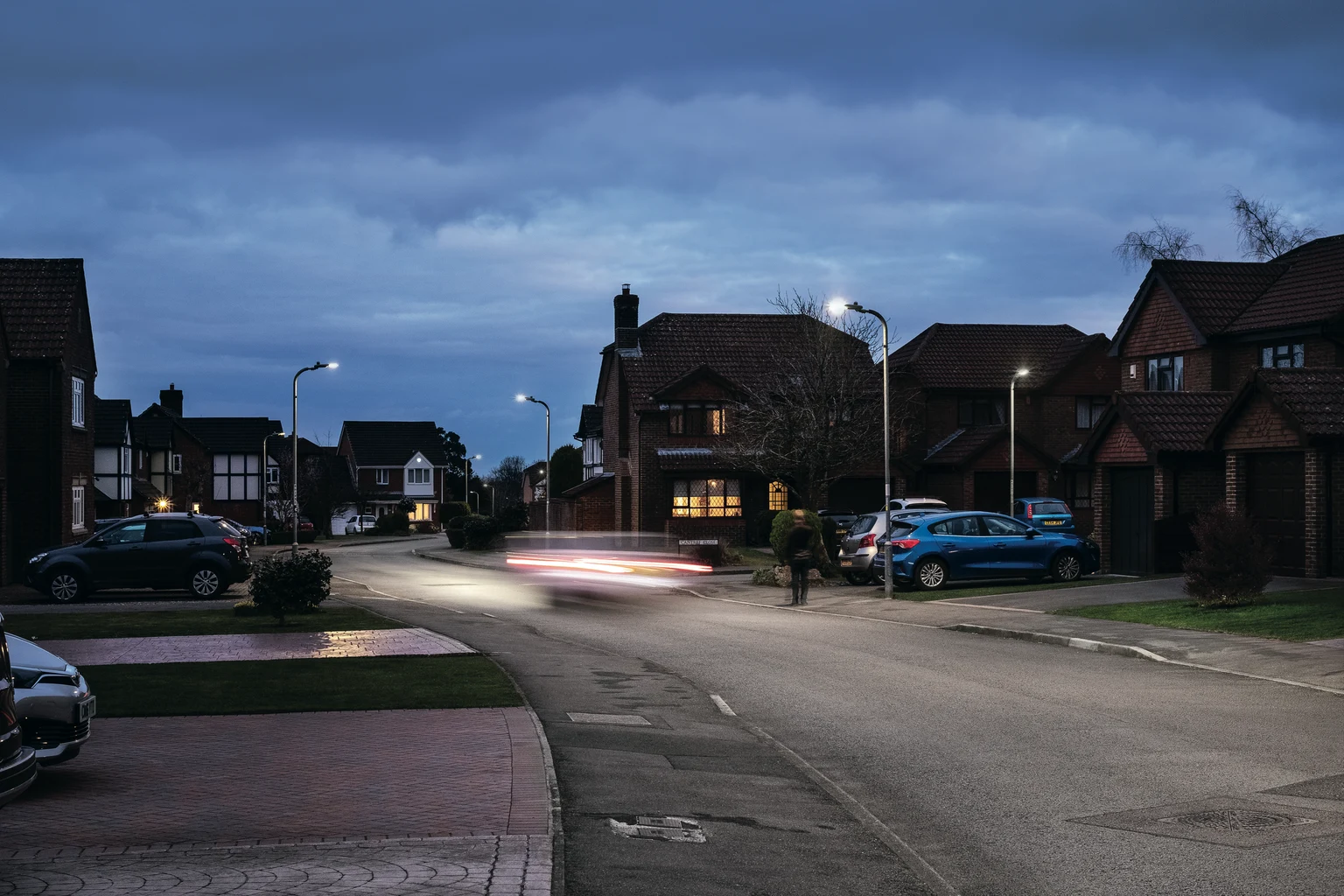 Eine ruhige Vorstadtstraße in der Abenddämmerung, mit Einfamilienhäusern und parkenden Autos auf beiden Seiten. Die Straßenleuchten sind eigeschaltet, und ein Auto fährt vorbei und hinterlässt Lichtspuren auf der Straße.