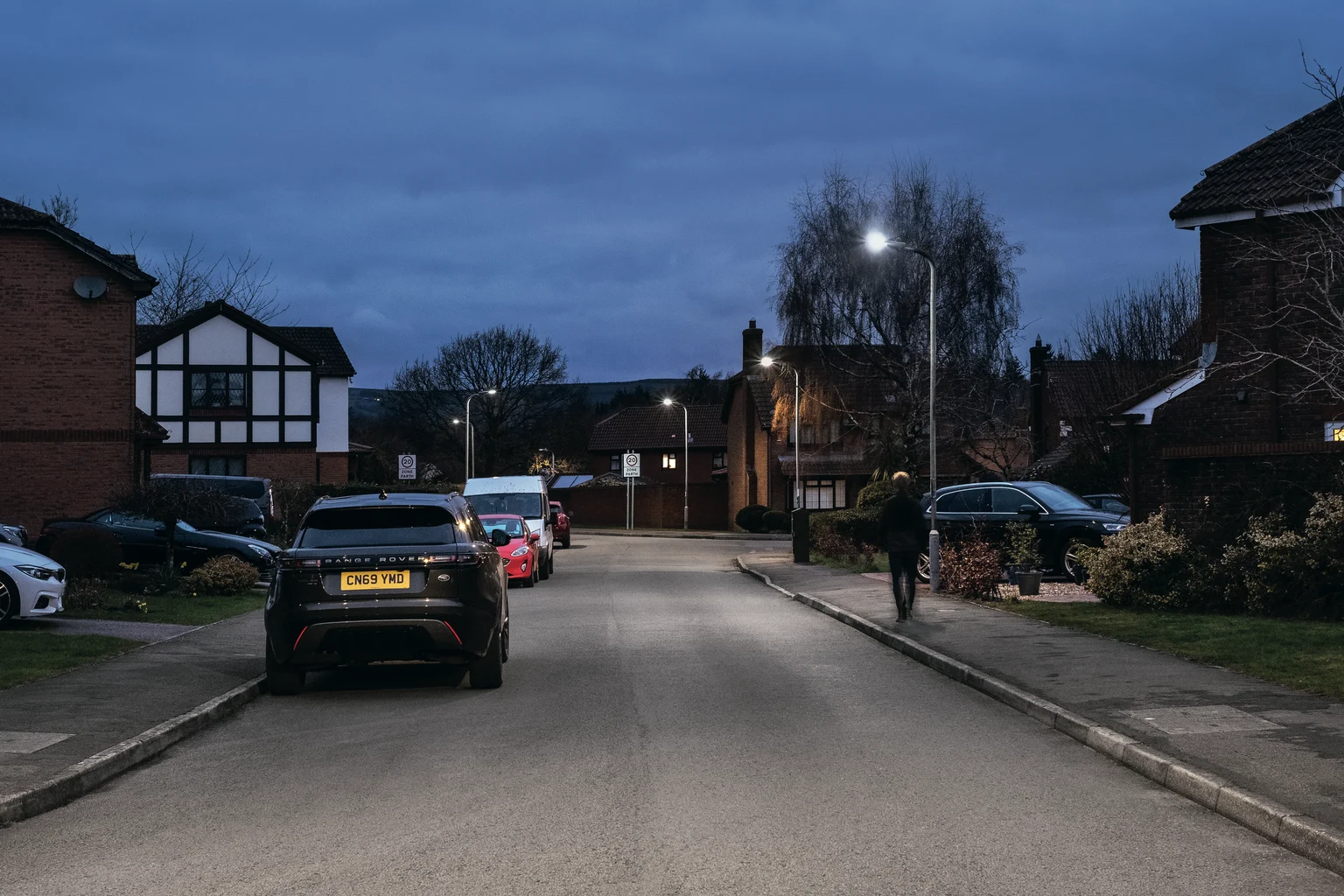 Eine ruhige Wohnstraße in der Abenddämmerung mit geparkten Autos auf beiden Seiten und freistehenden Häusern. Eine Person geht auf dem Bürgersteig unter eingeschalteten Straßenleuchten und einem bewölkten Himmel über ihr.