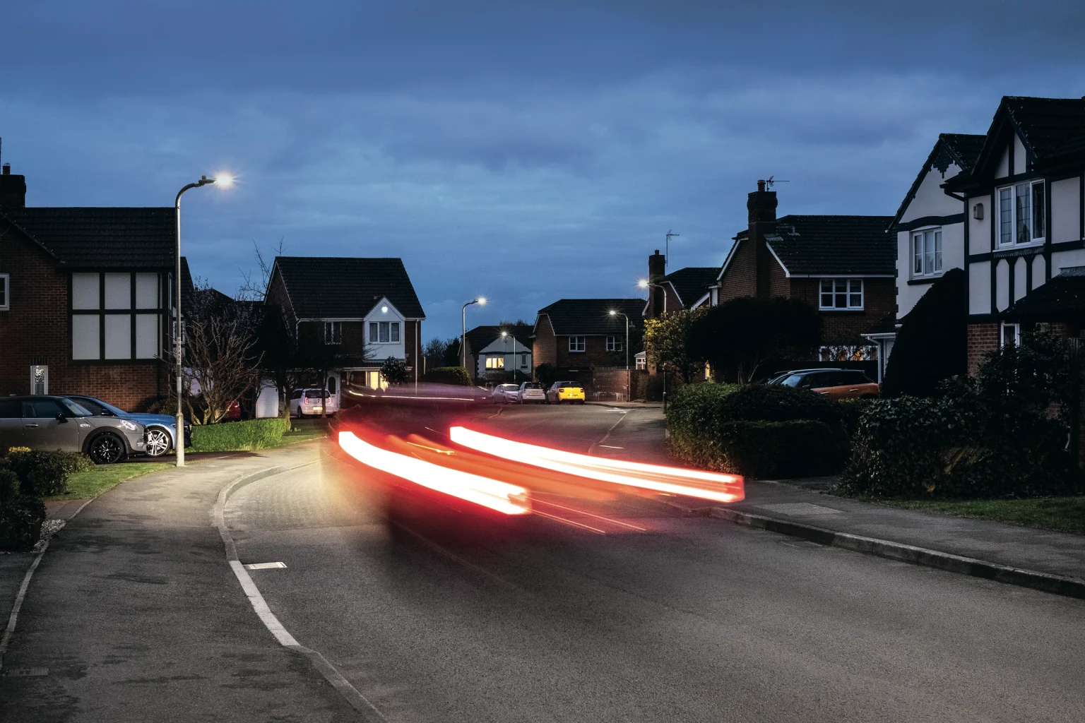 Eine ruhige Vorstadtstraße in der Abenddämmerung mit Einfamilienhäusern und geparkten Autos auf beiden Seiten. Die roten Lichtspuren eines vorbeifahrenden Autos sind sichtbar, und Straßenleuchten beleuchten die Szene unter einem bewölkten Himmel.