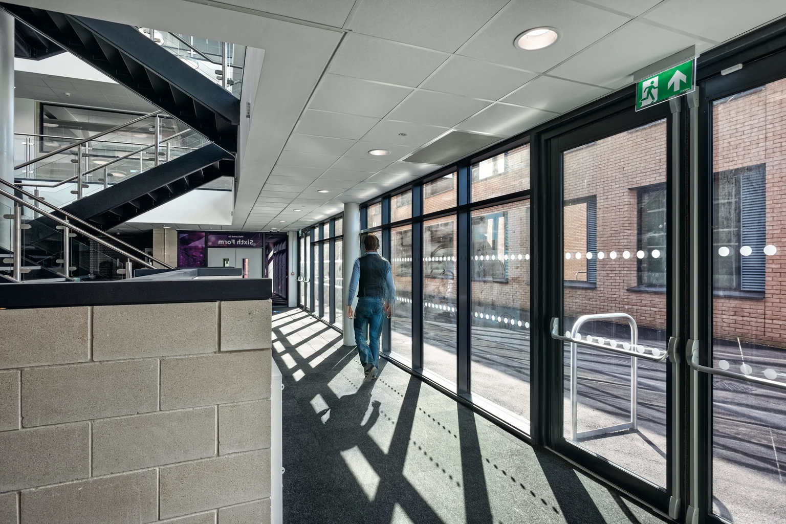 Corridoio luminoso con ampie finestre in vetro che permettono l'ingresso della luce naturale, completato da apparecchi illuminotecnici a soffitto incassati per un'illuminazione omogenea. Presenta elementi architettonici moderni, inclusa una scala e finiture lucide.