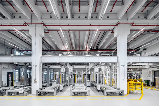 In einer hell ausgeleuchteten Industriehalle verlaufen mehrere Förderbänder zwischen weißen Stützsäulen und roten Rohrleitungen an der Decke.