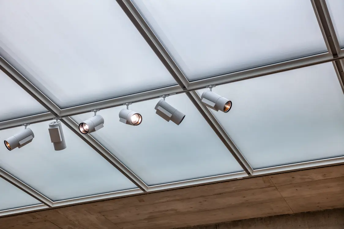 Een plafond met grote, matte glaspanelen die diffuus licht binnenlaten. Meerdere witte, cilindervormige spots zijn aan het plafond gemonteerd en zorgen voor gerichte verlichting.