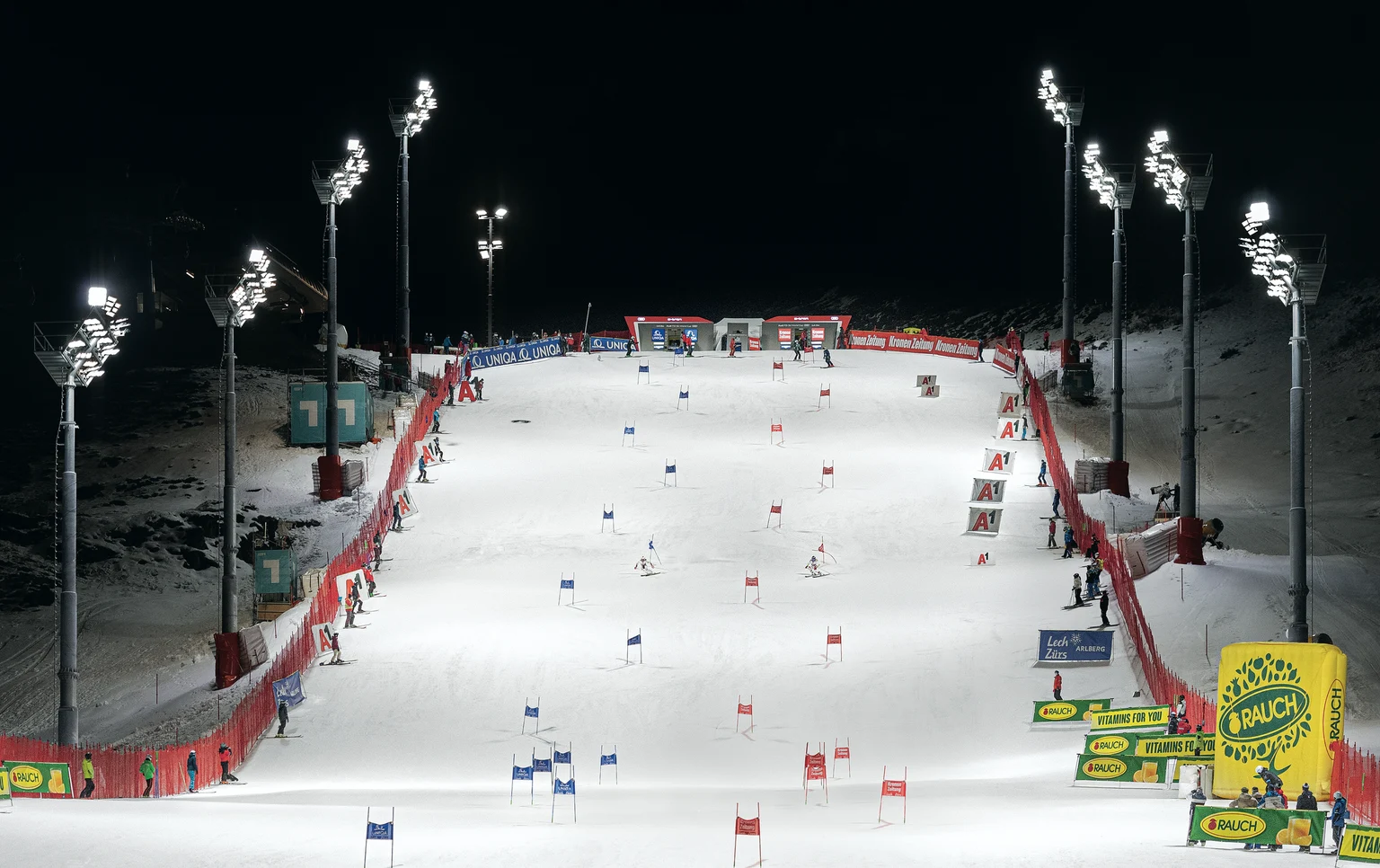 Une piste de ski illuminée la nuit avec plusieurs portes installées pour une compétition de slalom. Des mâts de projecteurs et des barrières de sécurité rouges ligne le parcours, tandis que des bannières publicitaires sont visibles dans la zone d'arrivée.