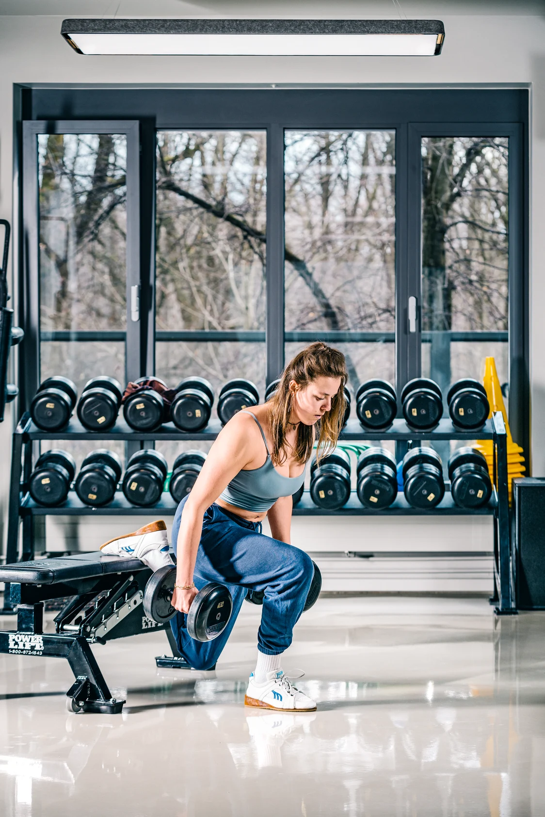 Il sistema di illuminazione TRAMAO illumina una sala fitness con luce naturale; una donna si allena con manubri davanti a una vetrata.