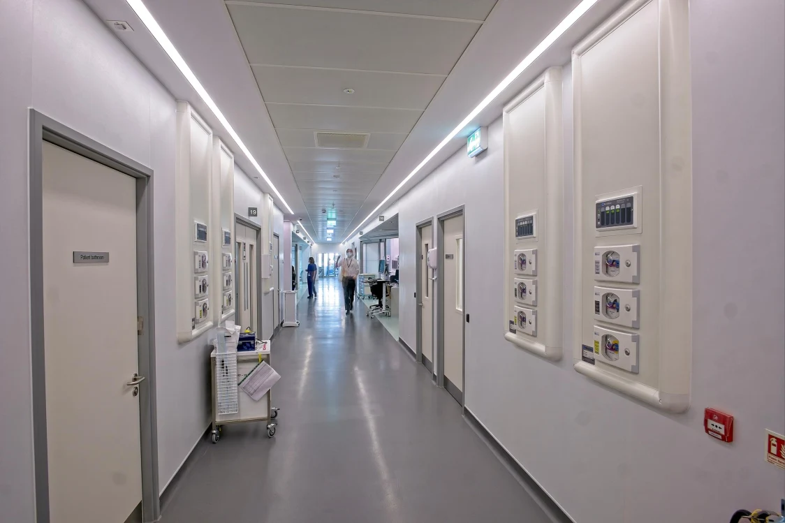 Un corridoio ospedaliero luminoso con pareti bianche e pavimento grigio, con porte e apparecchiature mediche visibili su entrambi i lati. Sullo sfondo diverse persone camminano lungo il corridoio.