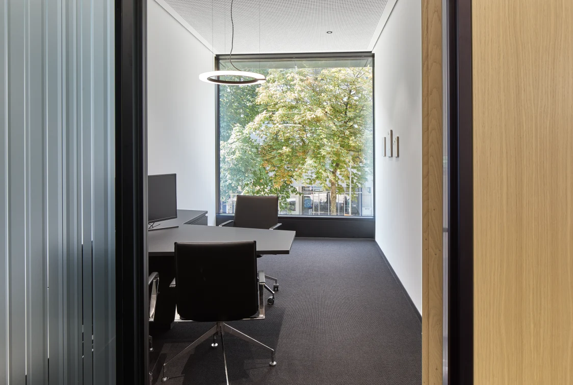 Moderner Besprechungsraum mit großem Fenster, ergonomischen Stühlen und einem schlichten Tisch. Eine ringförmige LED-Pendelleuchte sorgt für gleichmäßige Beleuchtung und eine klare Atmosphäre.