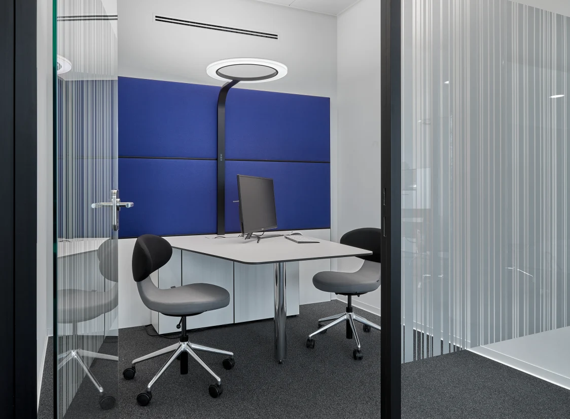 Kleiner Besprechungsraum mit blau gepolsterten Akustikwänden, modernem Tisch und ergonomischen Stühlen. Eine ringförmige LED-Leuchte sorgt für gleichmäßige Beleuchtung in der klar strukturierten Umgebung.