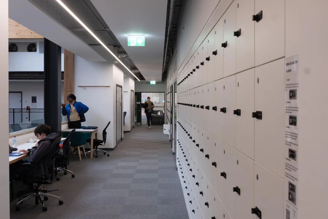 Un long couloir traverse une salle de lecture, bordée de casiers sur la droite. Un panneau de sortie de secours au plafond indique la direction.